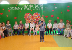 Chłopiec trzyma w ręku mikrofon, recytuje wiersz, w tle siedzą dzieci w kręgu.